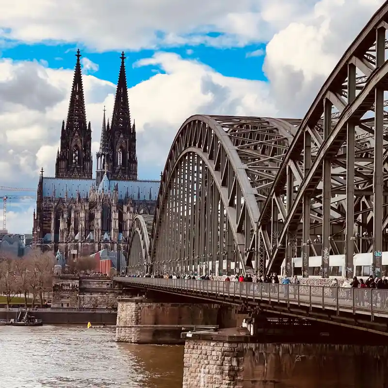 Der Kölner Dom hinter den Stahlbögen der Hohenzollernbrücke, auf dem Fußweg viele Menschen, unten der Rhein