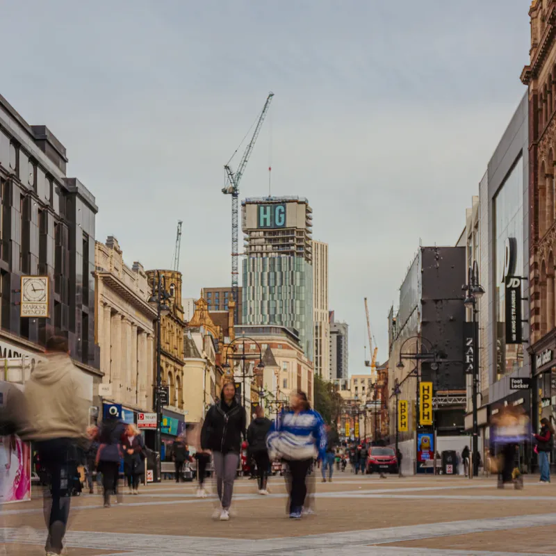 Shoppers walking down Briggate in Leeds city centre, with Victorian shopfronts and tower cranes against a grey sky