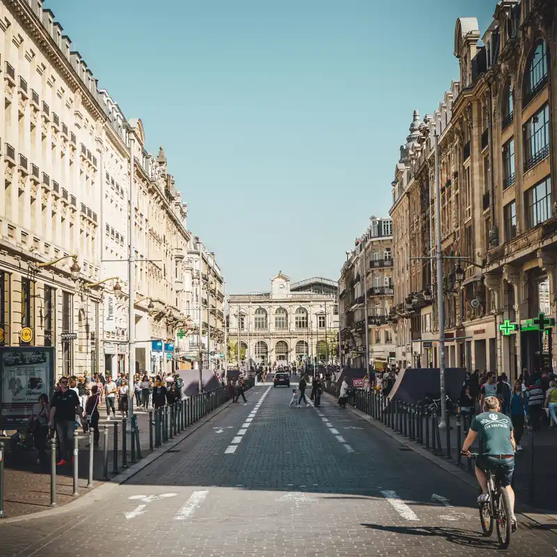 La rue Faidherbe, pavée et bordée d'immeubles haussmanniens, qui mène à la gare Lille-Flandres, avec une personne à vélo au premier plan et des piétons sur les trottoirs