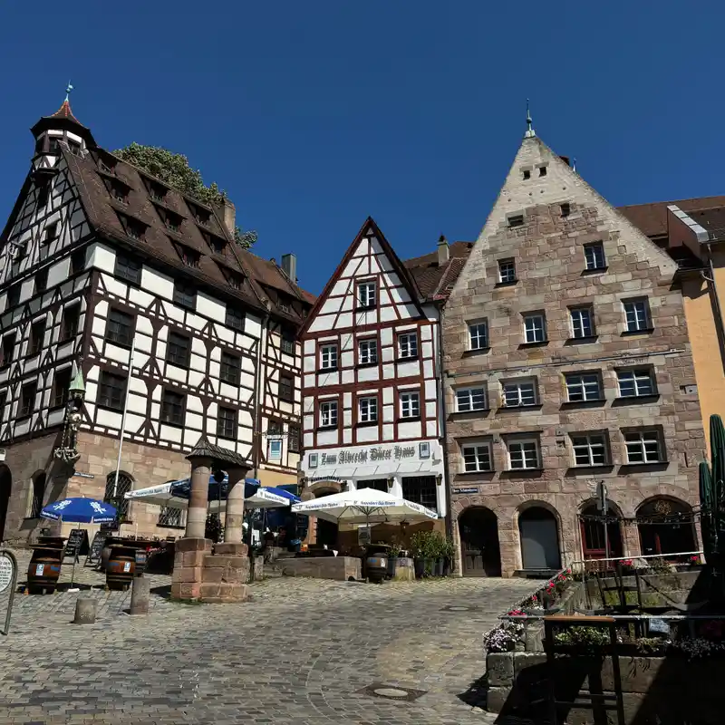 Fachwerkhäuser und ein Sandsteinhaus am Kopfsteinpflasterplatz in der Nürnberger Altstadt, davor Sonnenschirme eines Biergartens