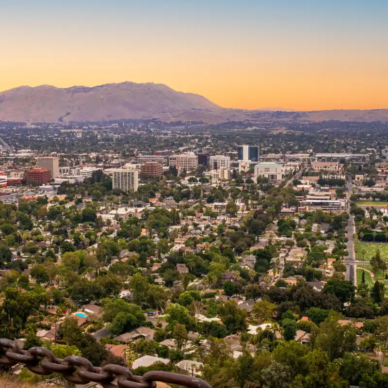 Downtown Riverside seen from a hillside at dusk, office towers rising above tree-lined streets, with bare mountains behind