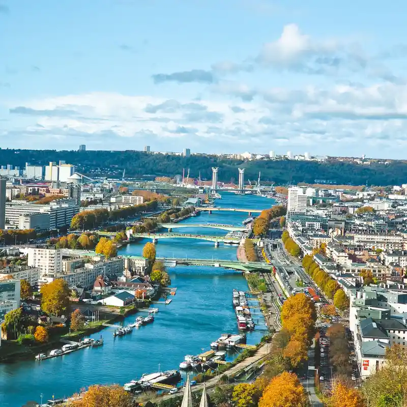 Vue aérienne de la Seine qui traverse Rouen en automne, avec ses ponts successifs, les quais bordés d'arbres et, au fond, les deux pylônes du pont Flaubert