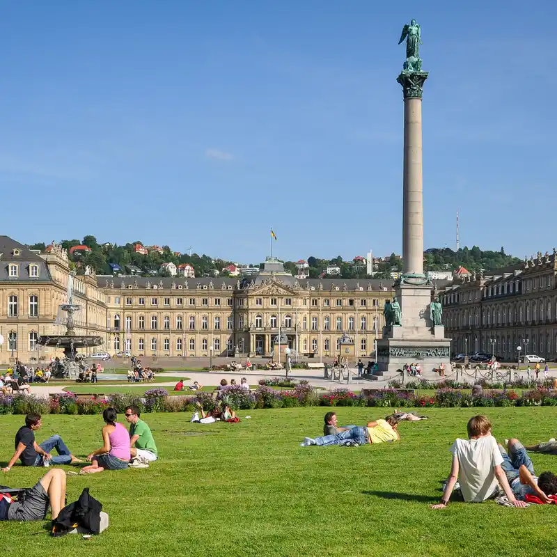 Menschen sitzen und liegen auf dem Rasen des Stuttgarter Schlossplatzes, dahinter das Neue Schloss mit Springbrunnen und rechts die Jubiläumssäule