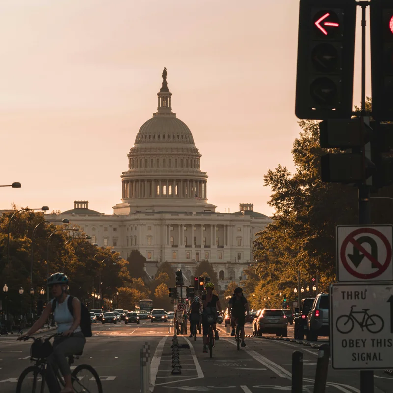 Cyclists riding a protected bike lane toward the United States Capitol at sunset, with a bike signal sign in the foreground