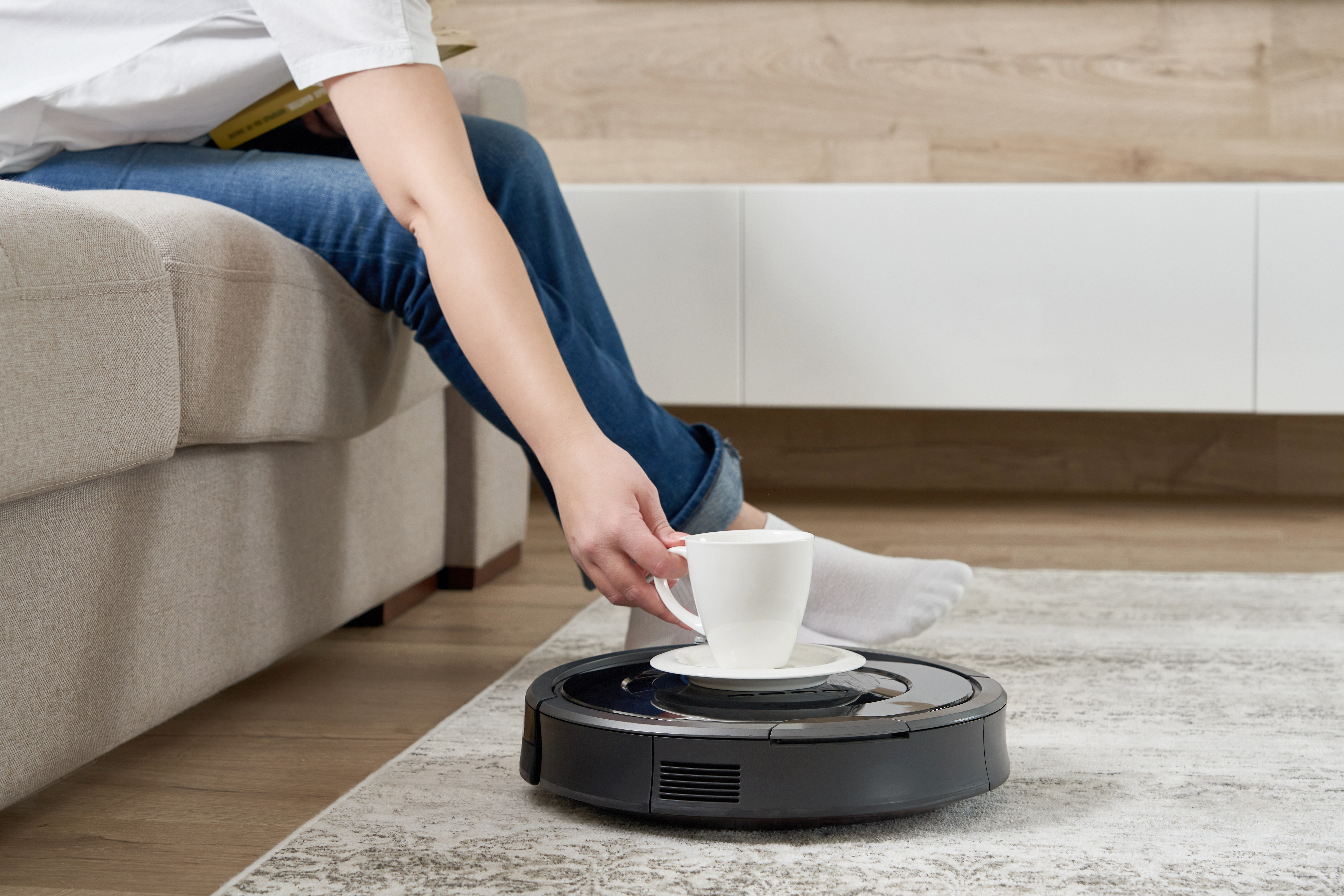 A robotic vacuum cleaner carries a tea cup across a living room floor. The vacuum cleaner is black and round, with two spinning brushes on the bottom. The tea cup is white. The vacuum cleaner is moving slowly and carefully, as if it is trying not to spill the tea. 